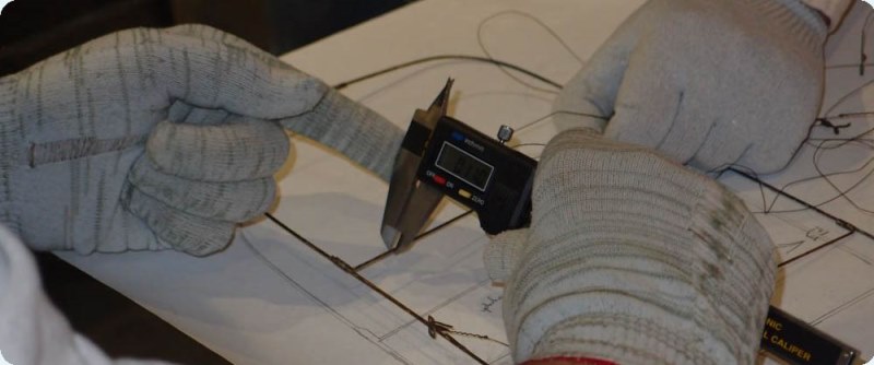 Two pairs of hands, both wearing white gloves, are measuring the diameter of a piece of metal making up the metal framework of a model airplane's wing.